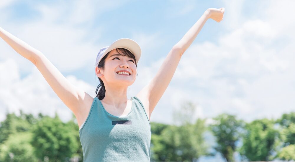 青空の下でキャップを被った女性が両腕を大きく広げて背伸びをしている画像で、【全身・胸郭(大胸筋・広背筋周辺)】を示し、姿勢改善や適切なケアによって呼吸が深くなり、心身ともにリフレッシュして活力に満ち溢れている様子。