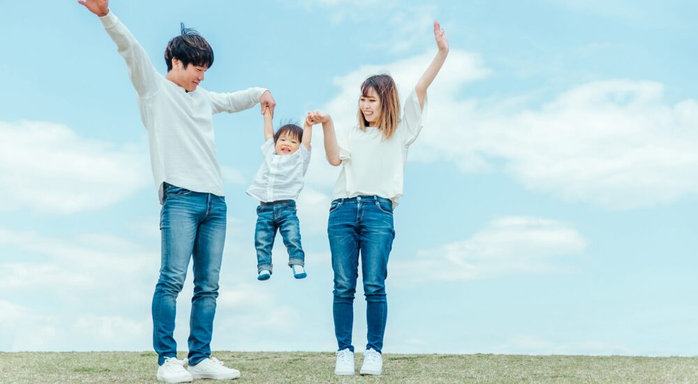 青空の下で両親が小さな息子の手を繋いで高く持ち上げ、笑顔で遊んでいる画像で、【全身・体幹(脊柱起立筋・骨盤周辺)】を示し、慢性痛から解放されることで心身に余裕が生まれ、家族との時間をアクティブに満喫している様子。