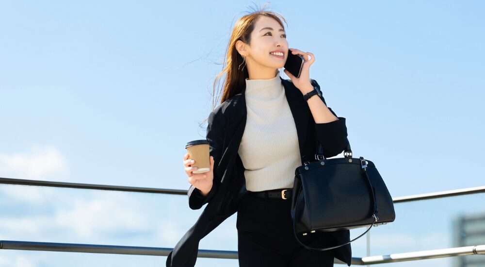 青空の下で女性がコーヒーとバッグを手に持ち、笑顔でスマートフォンで通話しながら颯爽と歩いている画像で、【体幹・下半身（大腰筋・骨盤周辺）】を示し、身体の軸が安定して片側に過度な負担をかけずに荷物を持てるようになり、エネルギッシュに通勤している様子。