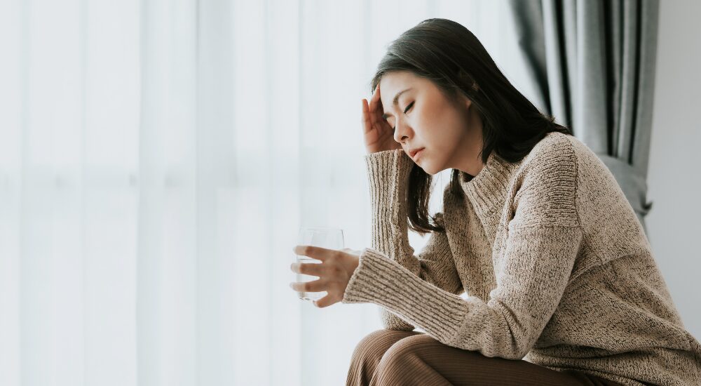 窓際でグラスを持ちながら、【頭部】を押さえ、頭痛やめまいなどの体調不良で辛そうにしている女性。