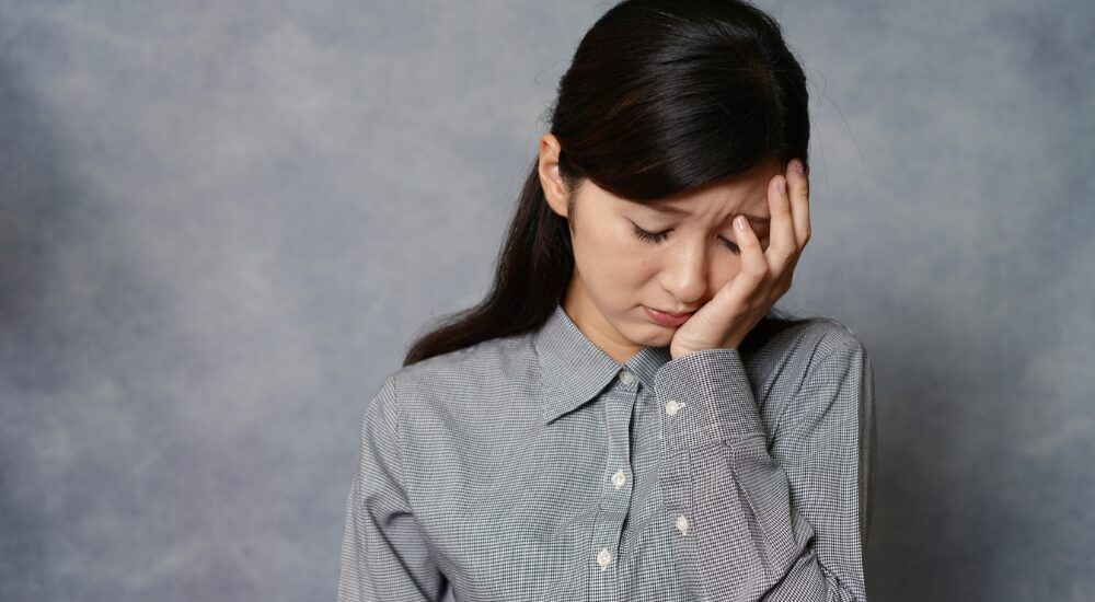 【側頭部(こめかみ)】を手で押さえ、深刻な頭痛や悩み事などで辛そうにうつむいているシャツ姿の女性。暗い雰囲気。