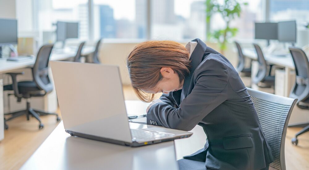 オフィスのデスクで、パソコンの前に突っ伏しているスーツ姿の女性。【心身（極度の過労・自律神経の失調）】により、仕事のストレスや睡眠不足が限界に達し、ぐったりとしている様子。