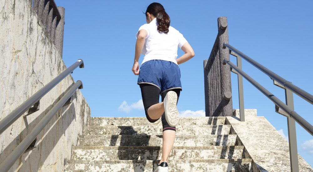 青空の下で女性が軽快に石段を駆け上がっている後ろ姿の画像で、【下半身・股関節（大臀筋・ハムストリングス周辺）】を示し、適切な身体の使い方や筋膜の滑走性向上により、足取りが軽くアクティブに運動を楽しめている様子。