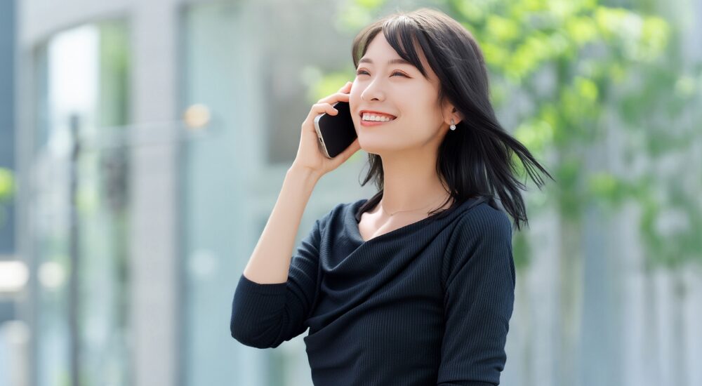 街の風景を背景に黒いトップスの女性が笑顔でスマートフォンで通話している画像で、店舗への問い合わせを示す画像。（CTAに使用可能）
